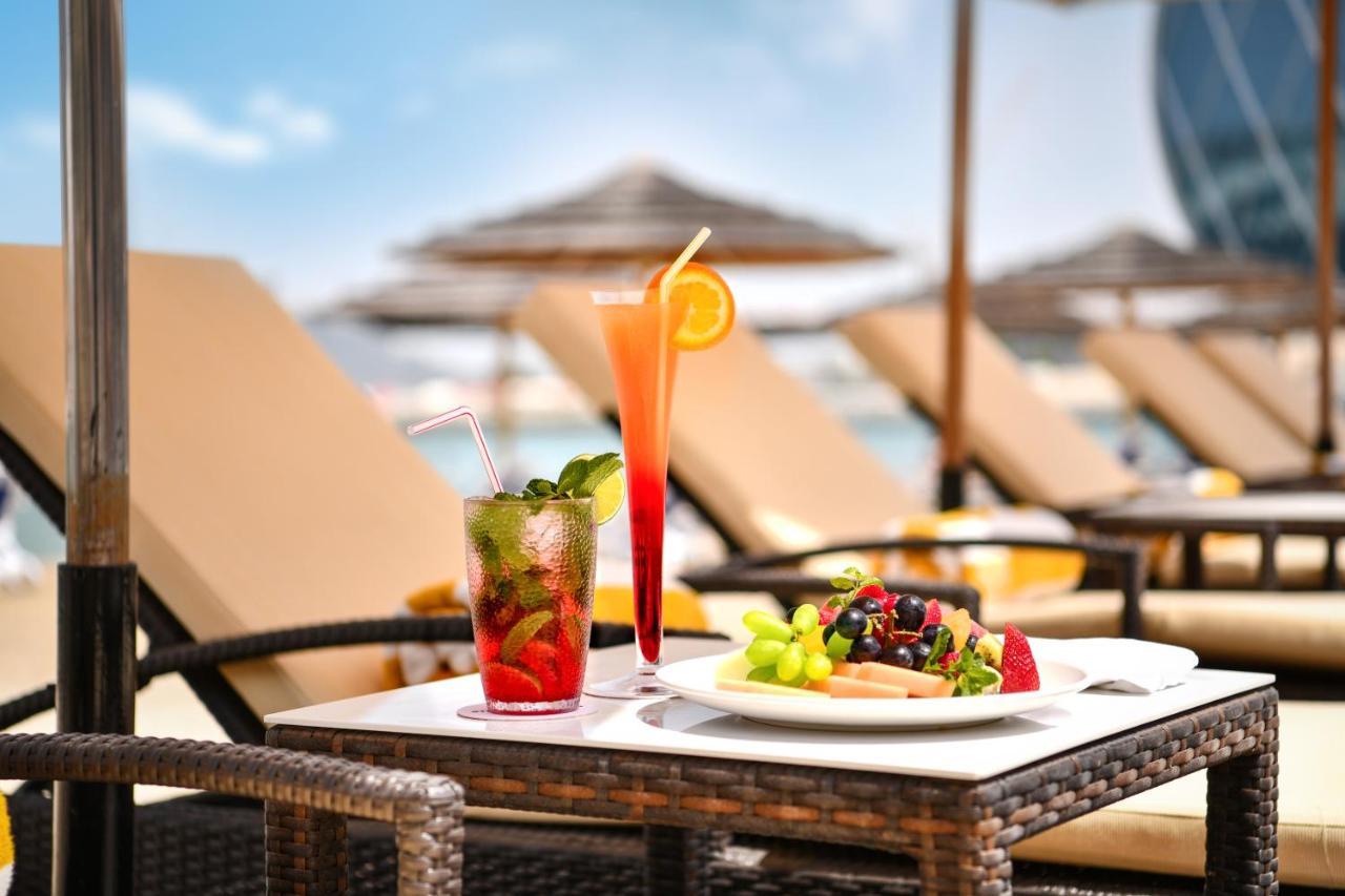 Al Raha Beach Resort & Spa Abu Dhabi Ngoại thất bức ảnh The photo shows a table set outdoors, featuring two colorful cocktails and a plate of food. One cocktail is a vibrant red drink garnished with mint, while the other is a layered orange beverage with an orange slice on the rim. The plate of food inclu