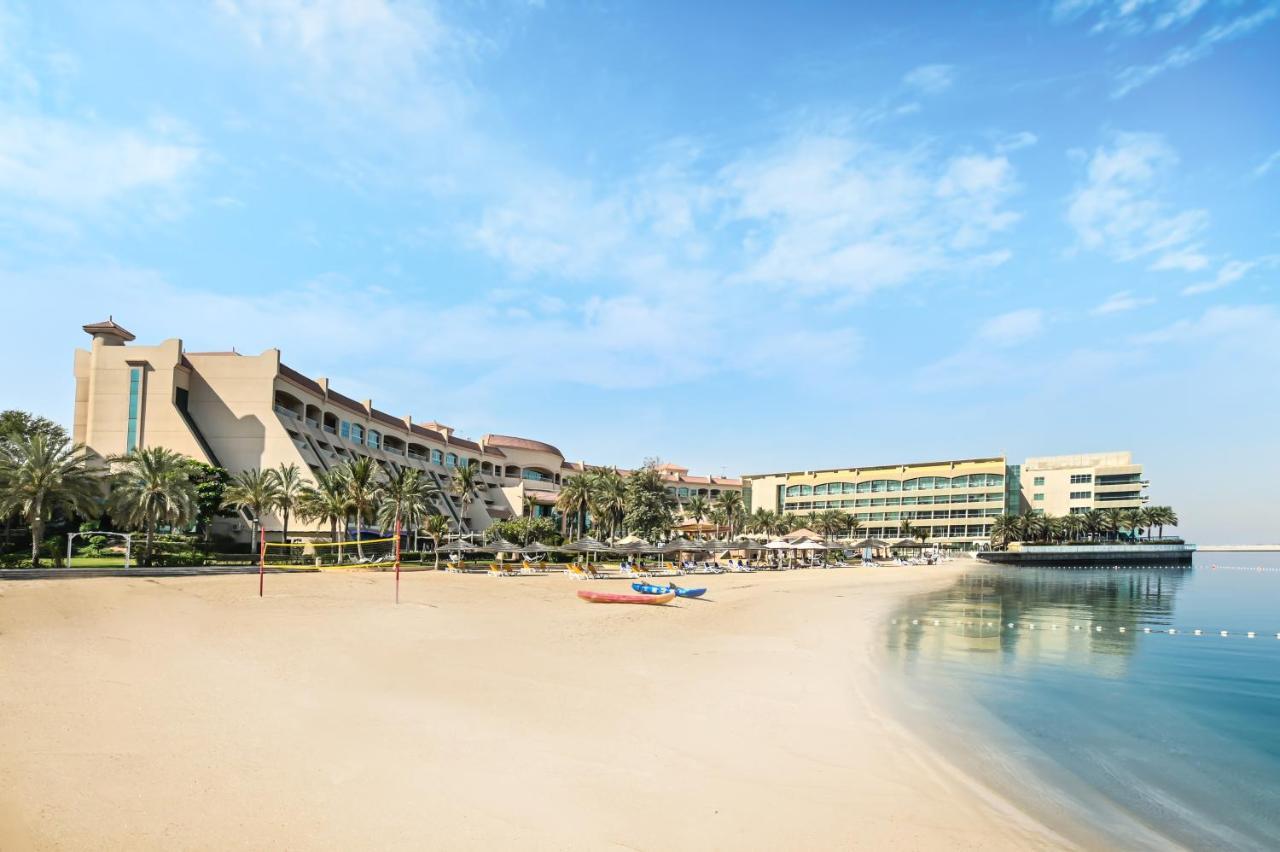 Al Raha Beach Resort & Spa Abu Dhabi Ngoại thất bức ảnh The photo depicts a serene beach scene with a clear blue sky. In the foreground, there is a sandy beach that gently slopes down to calm water. On the left side, several palm trees provide a tropical atmosphere. In the background, there are two large 