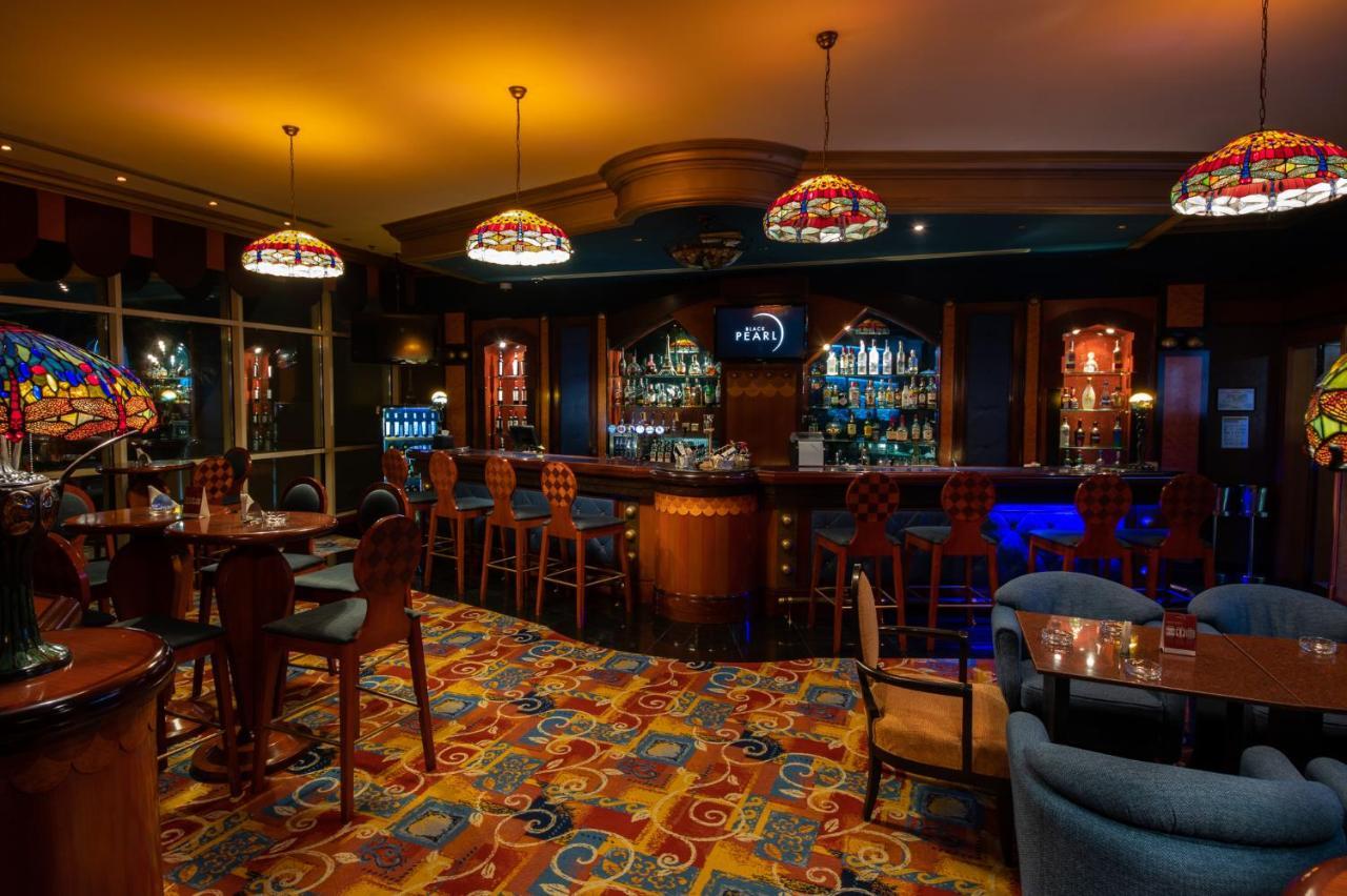 Al Raha Beach Resort & Spa Abu Dhabi Ngoại thất bức ảnh The photo shows a cozy bar interior, featuring a well-stocked bar area with various bottles displayed on shelves. The bar is illuminated by colorful stained glass light fixtures, creating a warm and inviting atmosphere. The floor is covered with a vi