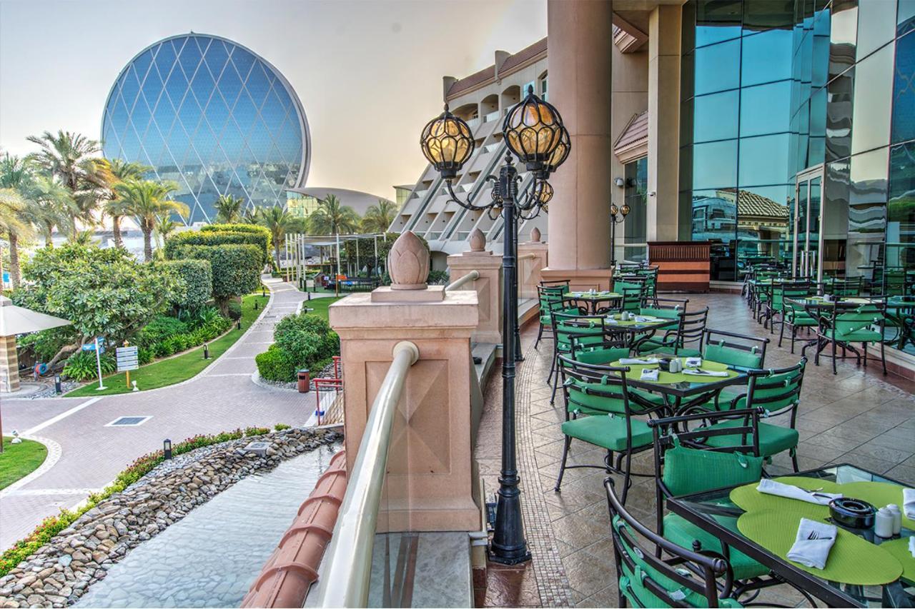 Al Raha Beach Resort & Spa Abu Dhabi Ngoại thất bức ảnh The photo depicts an outdoor dining area with green tables and chairs arranged on a patio. In the background, there is a large, distinctive building with a striking blue dome, surrounded by well-maintained landscaping, including trees and plants. The