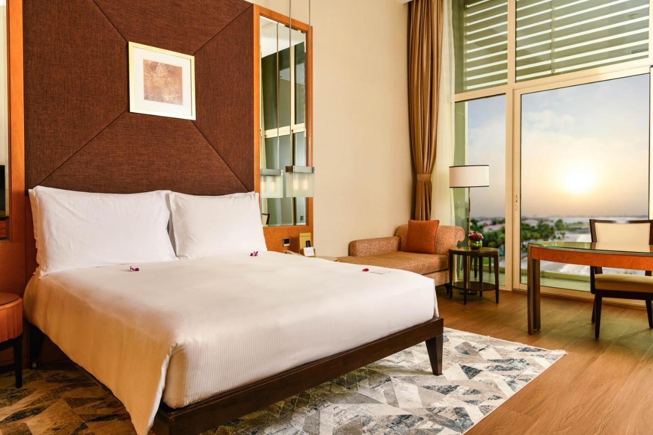 Al Raha Beach Resort & Spa Abu Dhabi Ngoại thất bức ảnh The photo shows a stylish hotel room featuring a large bed with white bedding, positioned against a textured wall. There’s a small seating area with a chair and a table, and a window with curtains that allows natural light to fill the space. The room