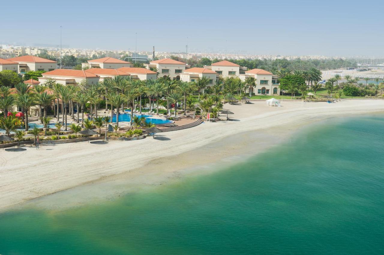 Al Raha Beach Resort & Spa Abu Dhabi Ngoại thất bức ảnh The photo depicts a serene beach scene with a clear turquoise sea and a sandy shoreline. In the background, there are several buildings featuring a Mediterranean architectural style with red-tiled roofs. The area is adorned with palm trees and outdoo