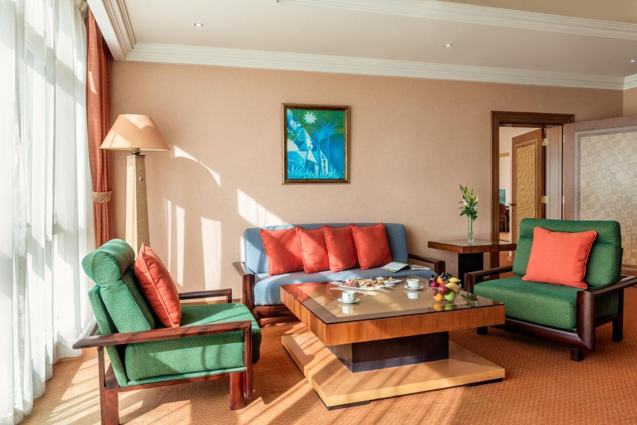 Al Raha Beach Resort & Spa Abu Dhabi Ngoại thất bức ảnh The photo shows a cozy living room with a warm ambiance. There are two sofas: one in green with red cushions and another in blue with red cushions, arranged around a wooden coffee table. On the table, there are some fruits and a small decorative item