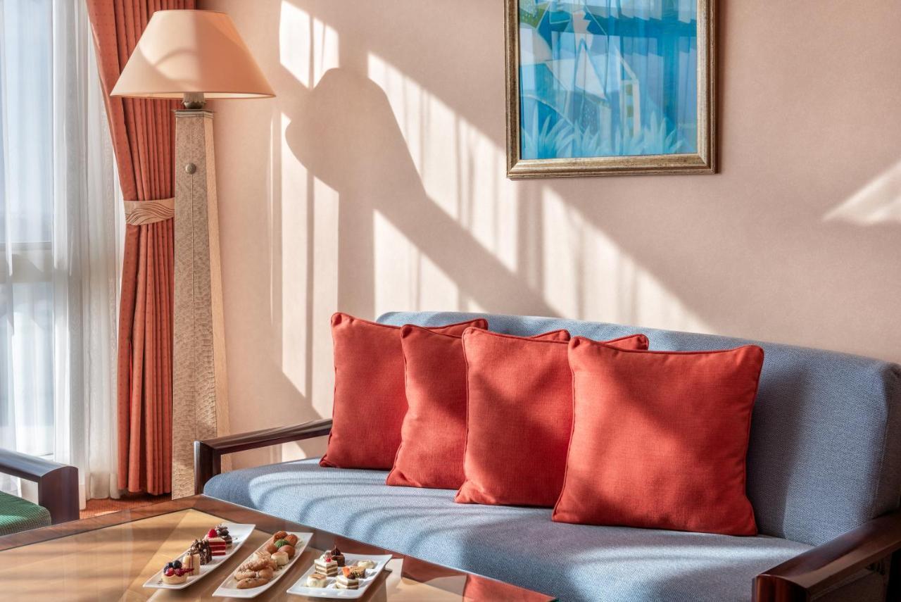 Al Raha Beach Resort & Spa Abu Dhabi Ngoại thất bức ảnh The photo shows a cozy interior setting, likely a sitting area. It features a light blue couch with four red cushions. In front of the couch, there is a glass table displaying an assortment of snacks or small dishes. The room has soft lighting, with 