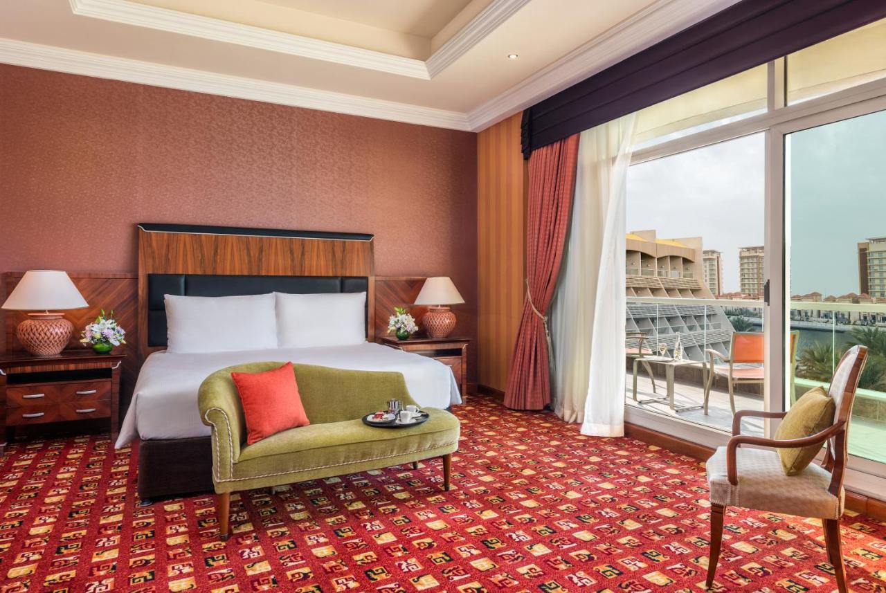 Al Raha Beach Resort & Spa Abu Dhabi Ngoại thất bức ảnh The photo shows a hotel room with an elegant and modern design. It features a large bed with white linens and a dark wooden headboard. On either side of the bed are matching bedside lamps. There is a green armchair with a red pillow positioned near t