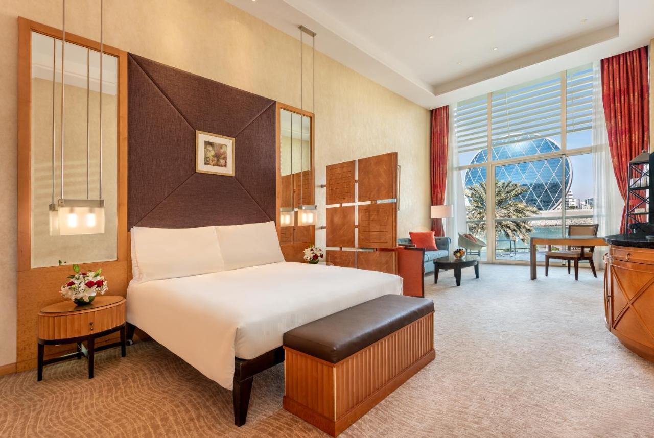 Al Raha Beach Resort & Spa Abu Dhabi Ngoại thất bức ảnh The photo shows a modern hotel room featuring a neatly made bed with a white comforter, situated against a stylish headboard with a geometric design. There are bedside tables on either side of the bed, each with a small flower arrangement. A comforta