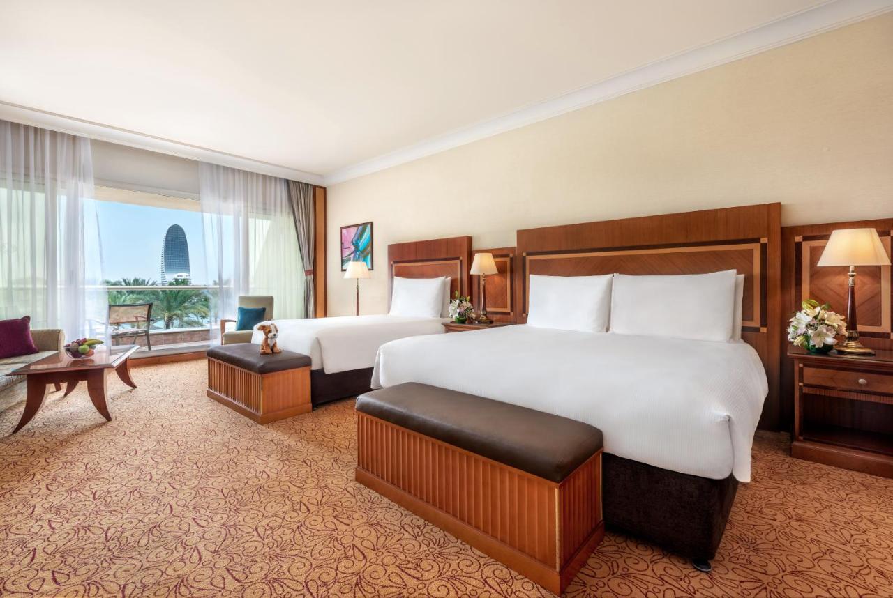 Al Raha Beach Resort & Spa Abu Dhabi Ngoại thất bức ảnh The photo shows a spacious hotel room featuring two large beds with white linens and dark brown upholstered headboards. There are matching benches at the foot of each bed. The room is decorated with a warm color palette and has a carpeted floor. To o
