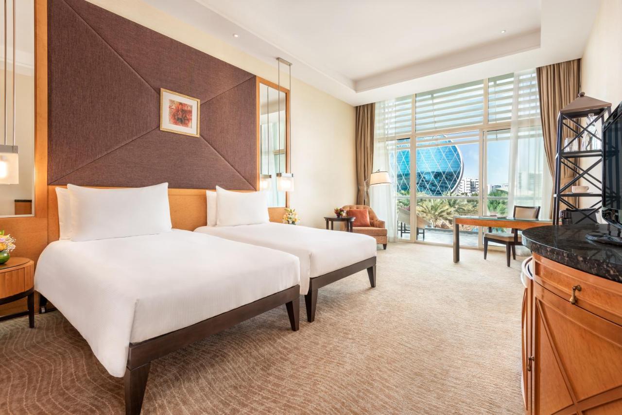 Al Raha Beach Resort & Spa Abu Dhabi Ngoại thất bức ảnh The photo shows a well-decorated hotel room featuring two single beds with white linens. The room has a modern aesthetic, with a large window letting in natural light and offering a view of a circular structure outside, possibly a landmark or artisti
