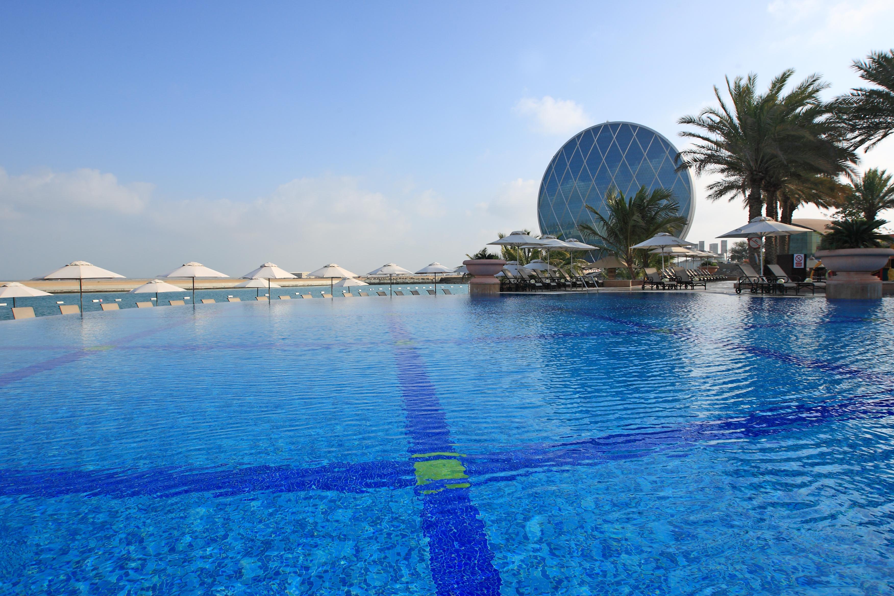 Al Raha Beach Resort & Spa Abu Dhabi Ngoại thất bức ảnh The photo features a large, serene swimming pool with clear, blue water reflecting the sky. Surrounding the pool are several lounge chairs and umbrellas, suggesting a relaxed, resort-like atmosphere. In the background, a distinctive building with a r