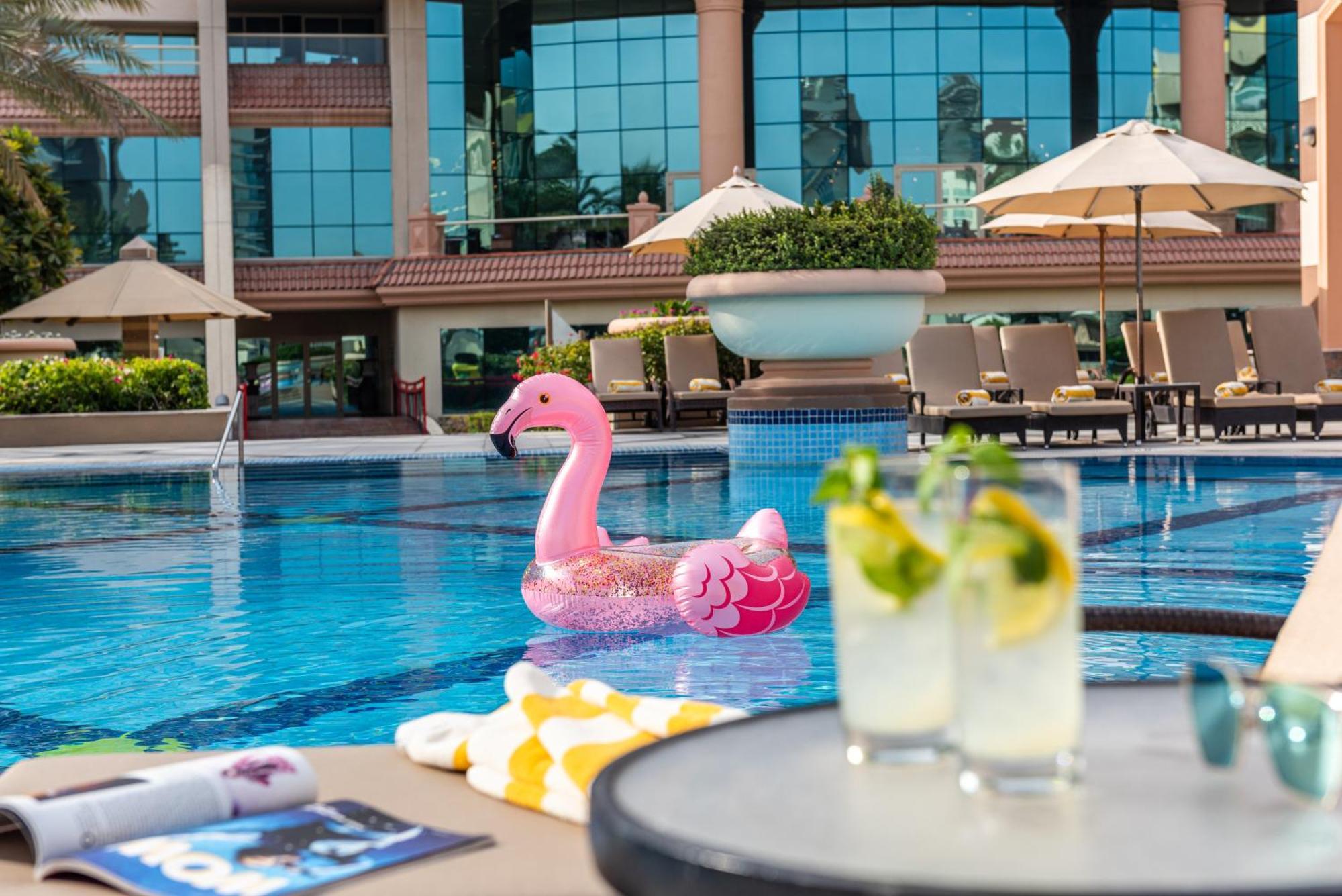 Al Raha Beach Resort & Spa Abu Dhabi Ngoại thất bức ảnh The photo depicts a bright and inviting outdoor swimming pool surrounded by a luxurious setting. In the pool, there is a large pink flamingo float, adding a fun and whimsical touch. To the side, a table is set up with two refreshing drinks, likely co