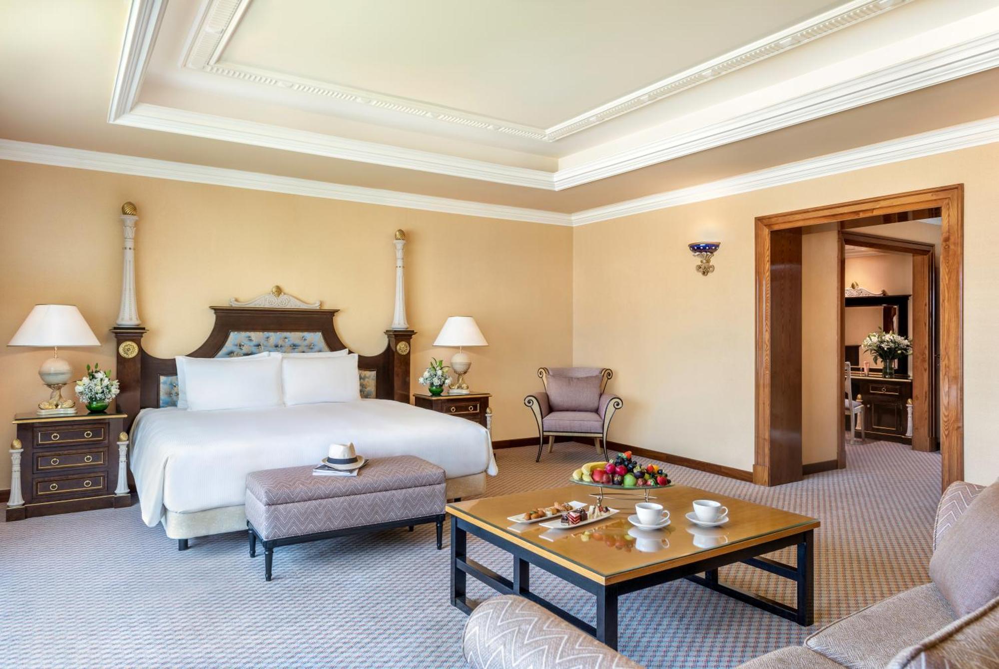 Al Raha Beach Resort & Spa Abu Dhabi Ngoại thất bức ảnh The photo shows an elegant hotel room, featuring a large bed with white linens, flanked by stylish bedside tables and lamps. The room has a warm color palette, with light brown walls and a plush carpet. A comfortable armchair is positioned nearby, an