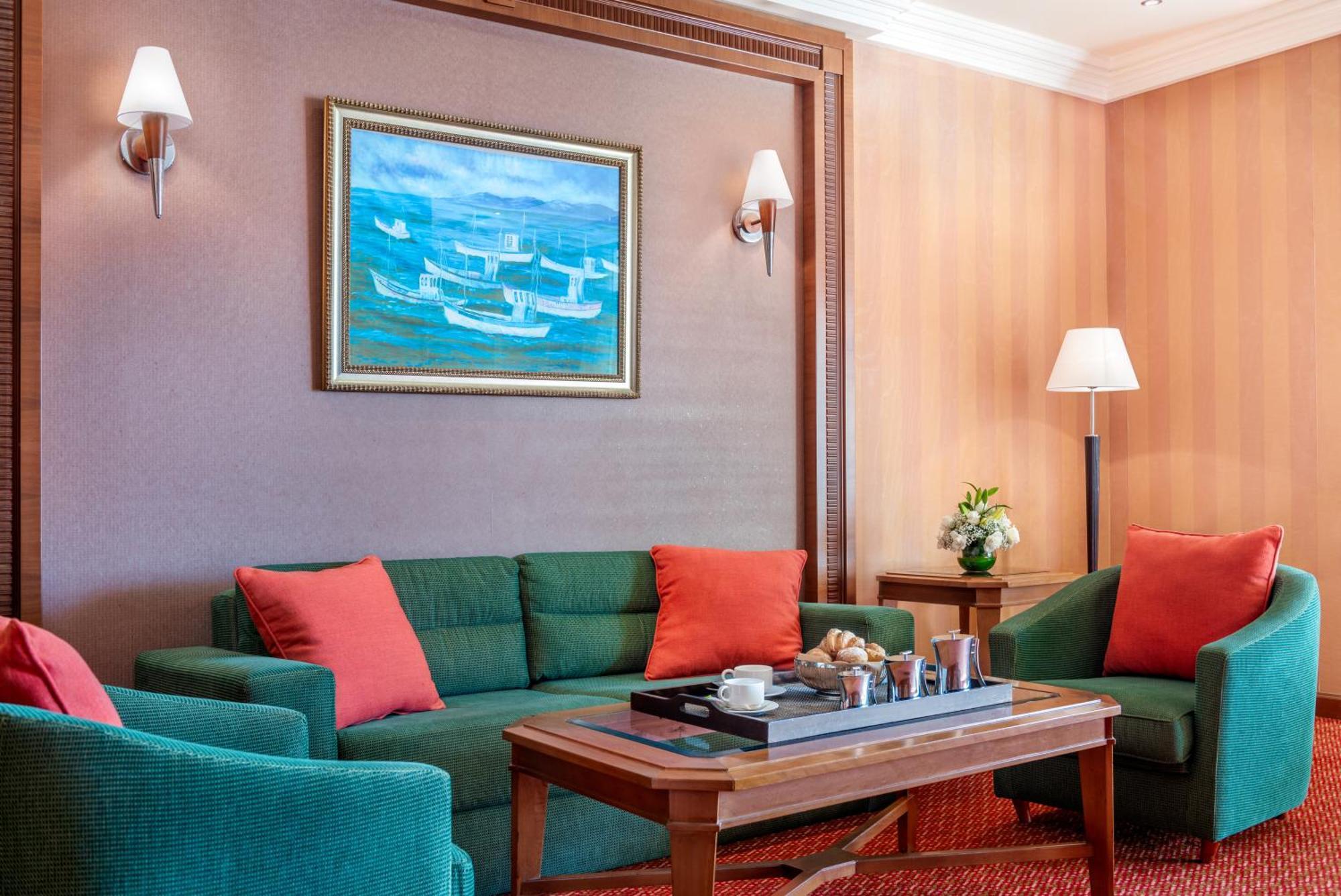 Al Raha Beach Resort & Spa Abu Dhabi Ngoại thất bức ảnh The photo displays a cozy and elegantly furnished living area. It features a green sofa adorned with orange cushions and two matching green armchairs. A wooden coffee table sits in the center, which holds a tray with beverages and snacks. The walls a