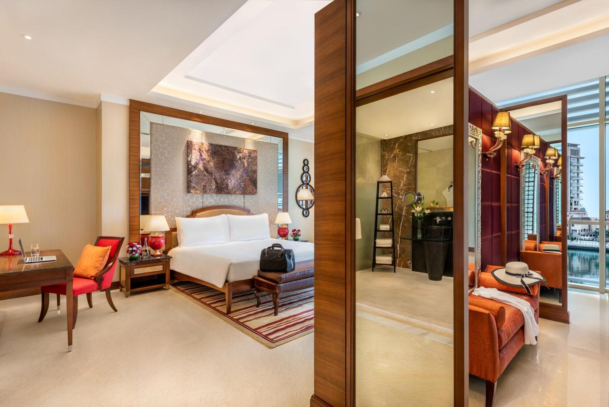 Al Raha Beach Resort & Spa Abu Dhabi Ngoại thất bức ảnh The photo shows a luxurious hotel room with a modern design. The room features a large bed with white linens, flanked by bedside tables with lamps. There is a area with a chair and a small table on the left side, and a vibrant artwork hanging above t