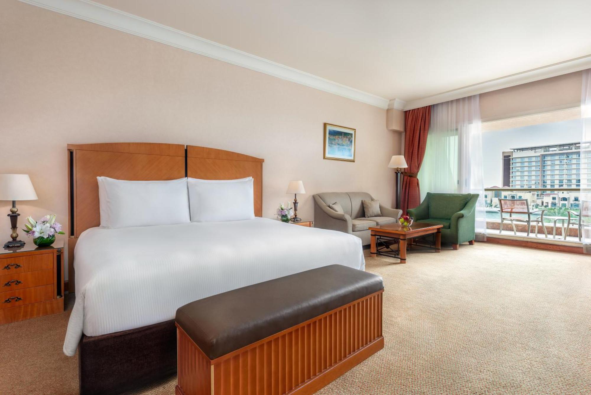 Al Raha Beach Resort & Spa Abu Dhabi Ngoại thất bức ảnh The photo shows a well-furnished hotel room featuring a large bed with white linens and a wooden headboard. There's a brown bench at the foot of the bed. To one side, a sitting area includes a sofa and a coffee table, complemented by two lamps. Large