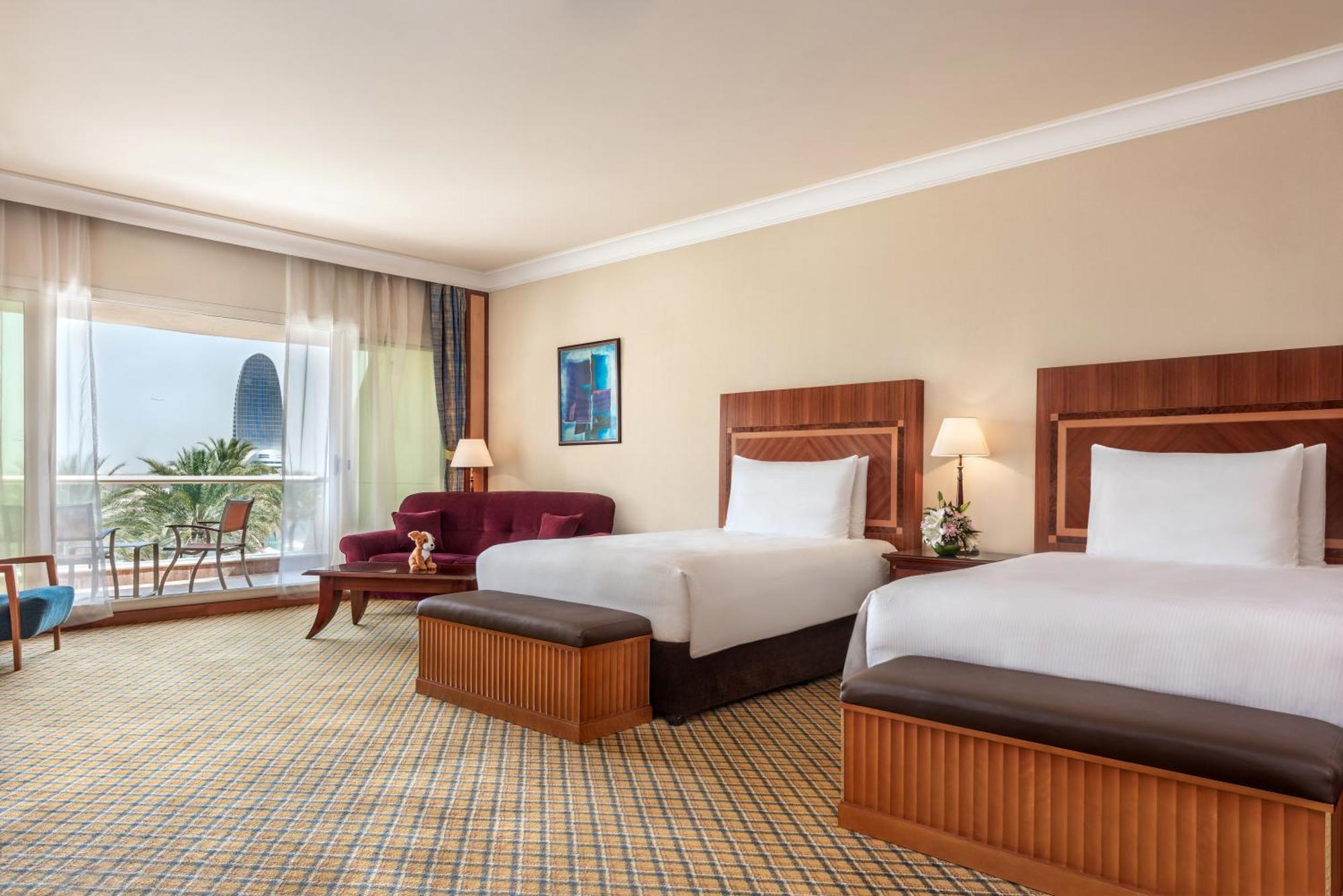 Al Raha Beach Resort & Spa Abu Dhabi Phòng bức ảnh The photo shows a hotel room featuring two twin beds with wooden headboards. The room has a light, neutral color scheme, with a large window that provides natural light and a view of the outdoors. There is a small seating area with a purple sofa and 