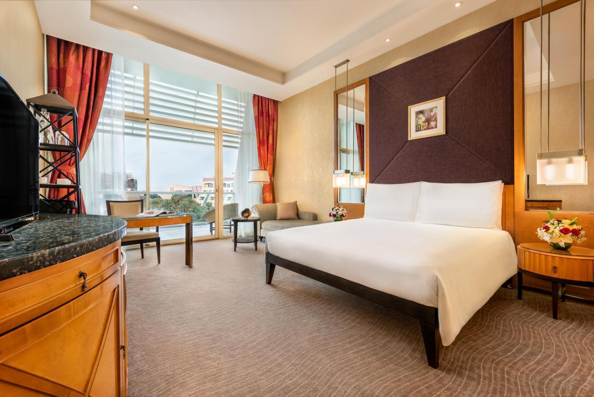 Al Raha Beach Resort & Spa Abu Dhabi Phòng bức ảnh The photo shows a modern hotel room featuring a large bed with a white duvet positioned against a purple accent wall. There are tall windows with sheer curtains allowing natural light to enter the space, along with red drapes framing the windows. A d