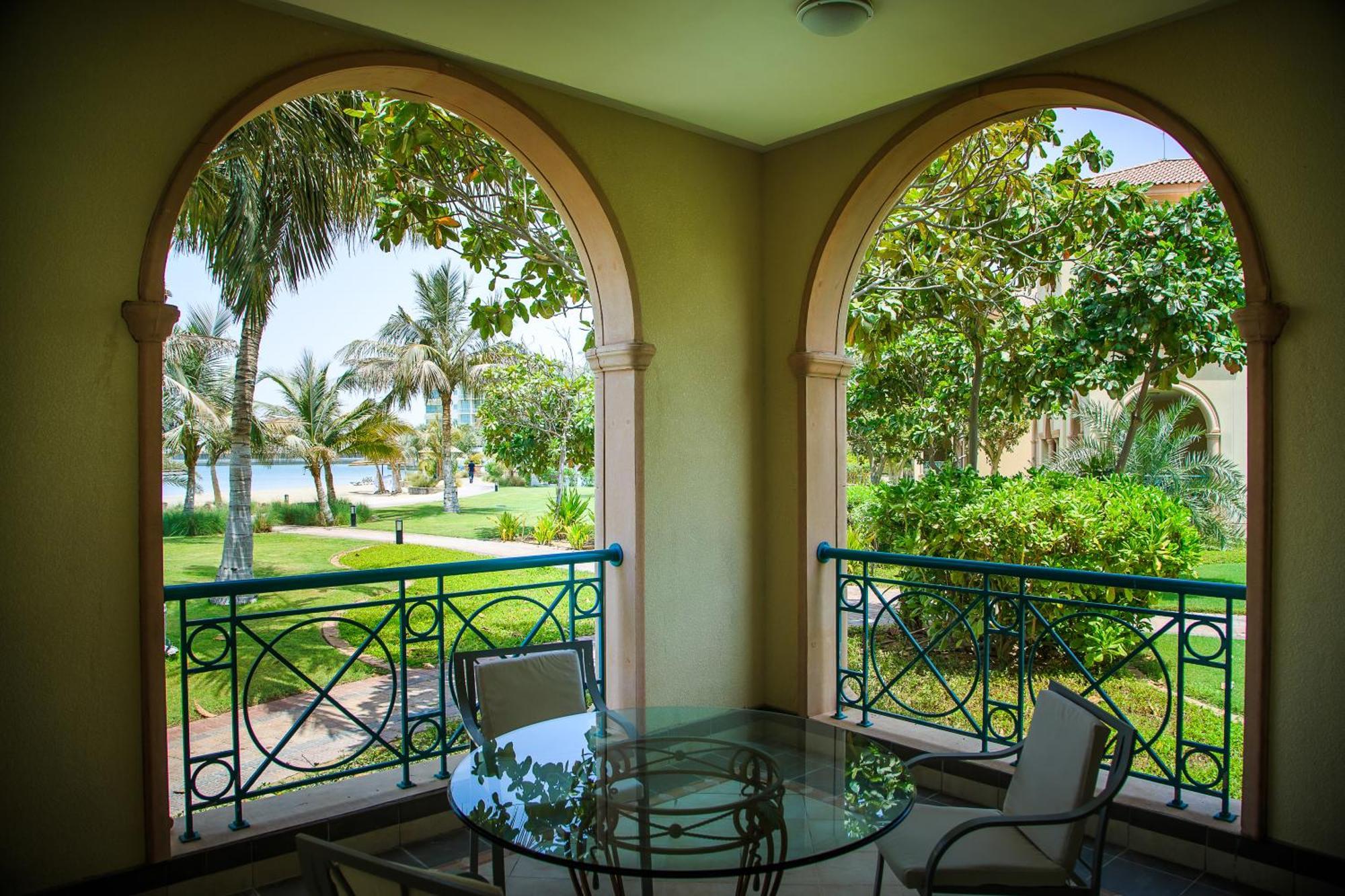 Al Raha Beach Resort & Spa Abu Dhabi Phòng bức ảnh The photo displays a beautifully designed balcony or terrace area. It features two large arches that frame the view outside. Through the arches, you can see a lush garden with palm trees and well-maintained greenery. There’s a glass table with severa