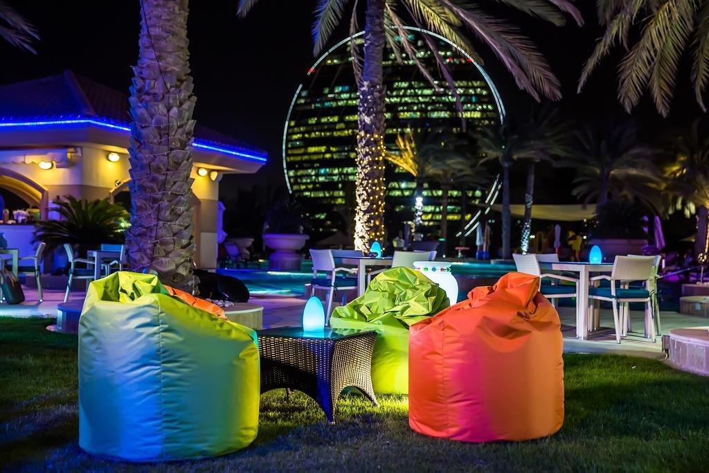 Al Raha Beach Resort & Spa Abu Dhabi Ngoại thất bức ảnh The photo shows a vibrant outdoor lounge area at night. In the foreground, there are colorful bean bag chairs—one in bright green, one in yellow, and one in orange. A small table sits between the bean bags. In the background, illuminated palm trees a