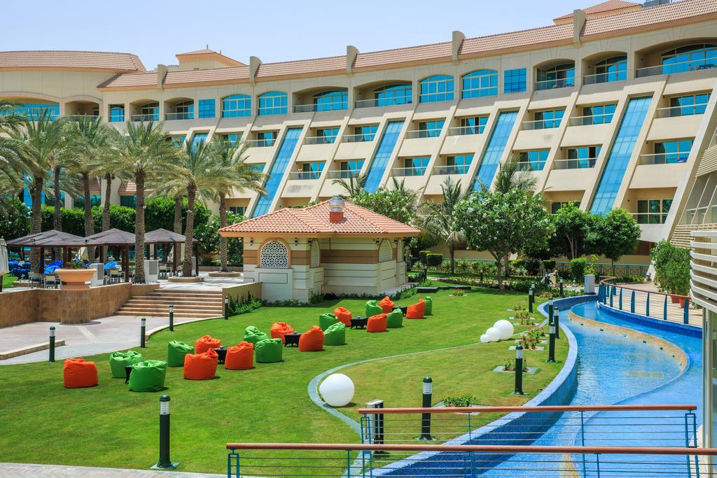 Al Raha Beach Resort & Spa Abu Dhabi Ngoại thất bức ảnh The photo depicts a modern hotel complex with multiple stories and large windows. In the foreground, there is a well-maintained lawn featuring colorful lounge chairs in shades of orange and green. A pathway runs alongside a blue water feature, creati