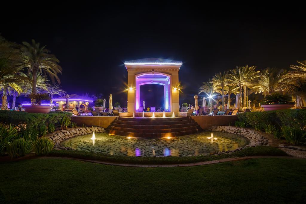 Al Raha Beach Resort & Spa Abu Dhabi Ngoại thất bức ảnh The photo depicts a beautifully lit outdoor scene at night. In the foreground, there is a serene water feature with a circular pond, and small fountains creating gentle ripples. Surrounding the pond are lush green lawns and ornamental plants. 

In th