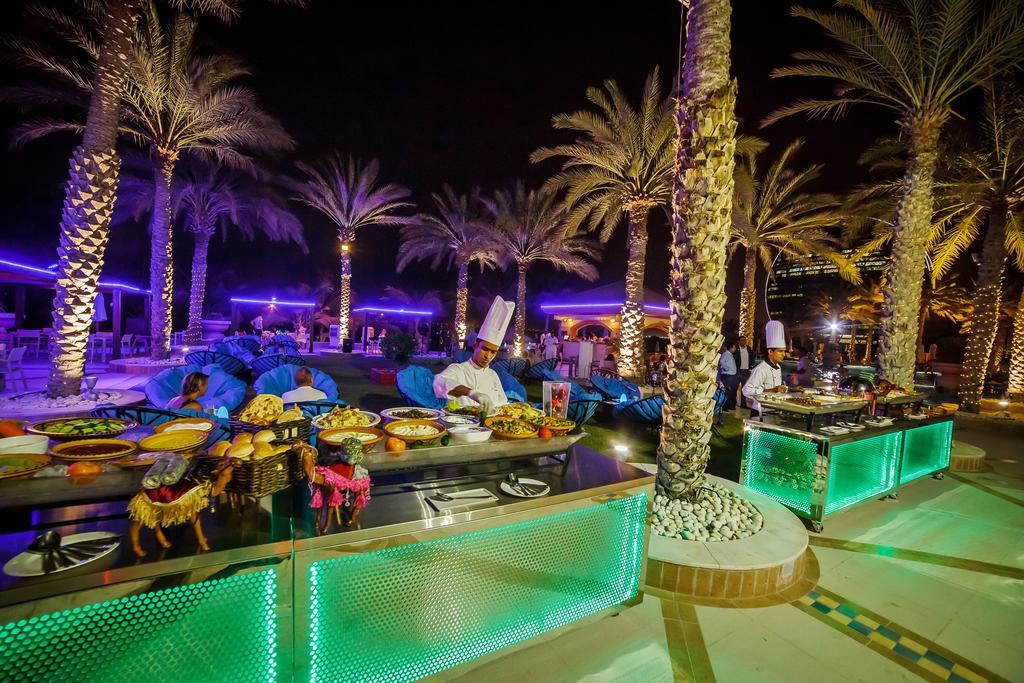 Al Raha Beach Resort & Spa Abu Dhabi Ngoại thất bức ảnh The photo depicts an outdoor dining area at night, featuring several chefs in white uniforms and hats working at a food station. The setting is illuminated by colorful lights, with glowing green accents on the serving tables. Surrounding the area are